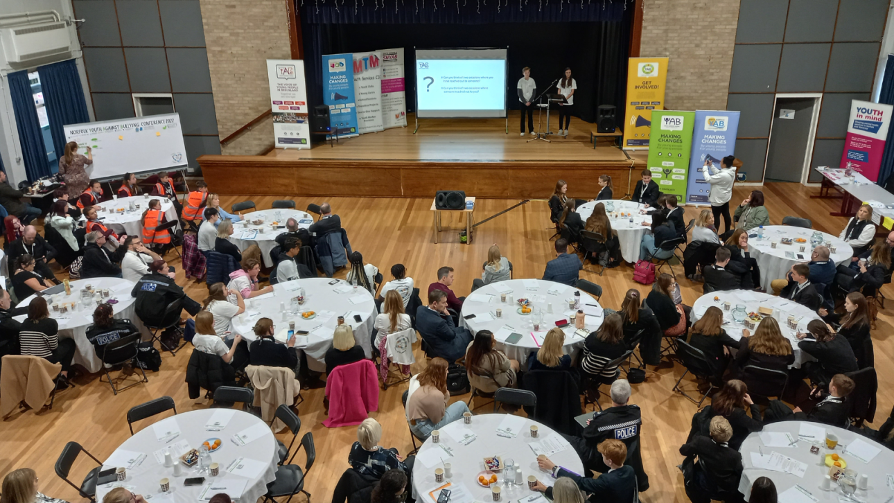 Large hall of people sat at tables watchnig a presentation from last year's conference