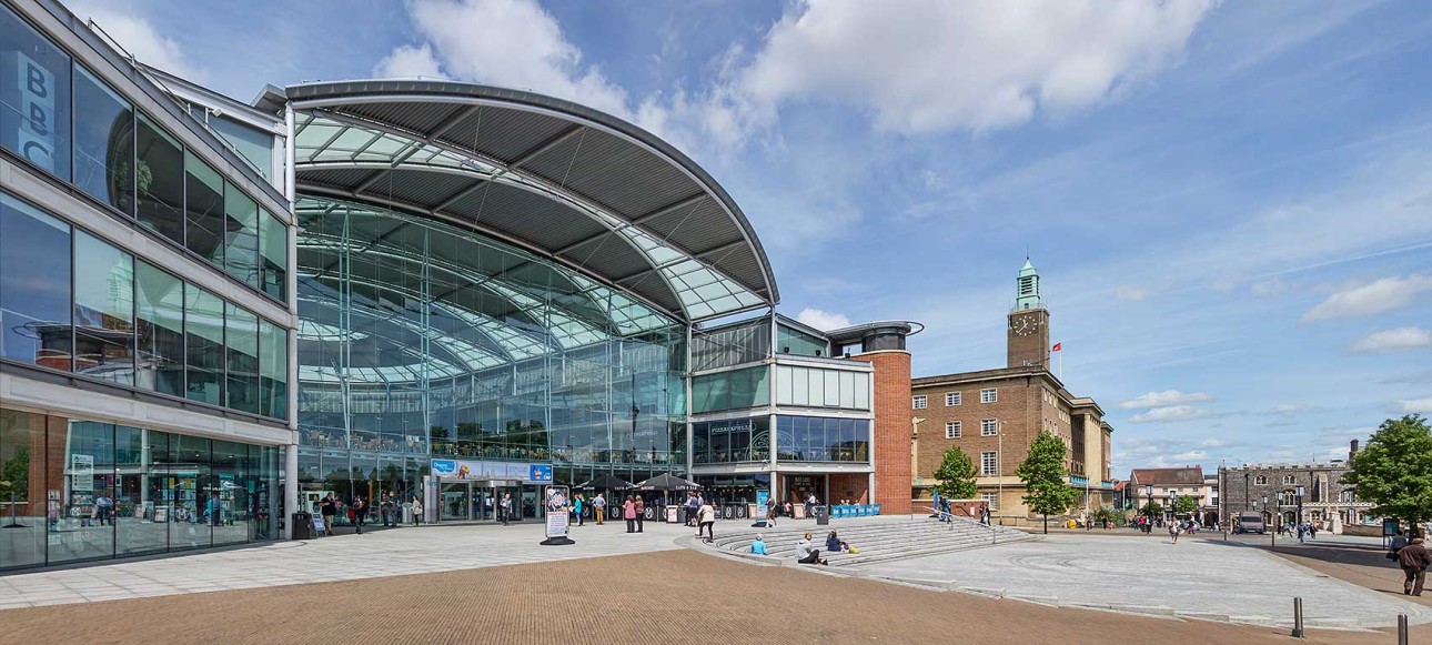 The exterior of the Forum in Norwich on a sunny day, viewed with the Council building and clock tower visible in the distance.