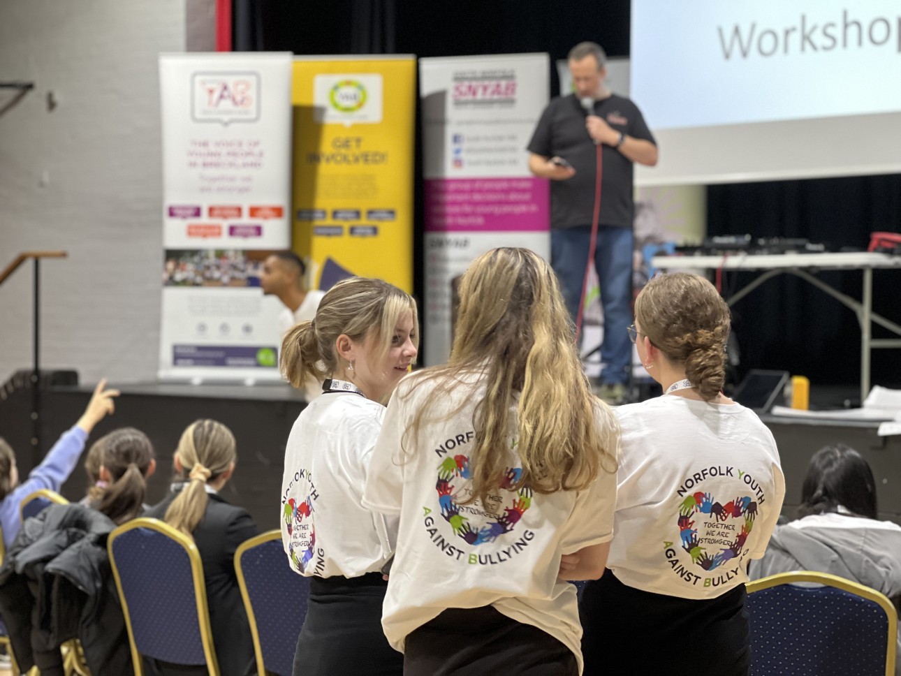 A trio of young people in branded NYAB tee shirts talk amongst themselves, whilst a speaker stands on stage behind them with a microphone, taking questions.