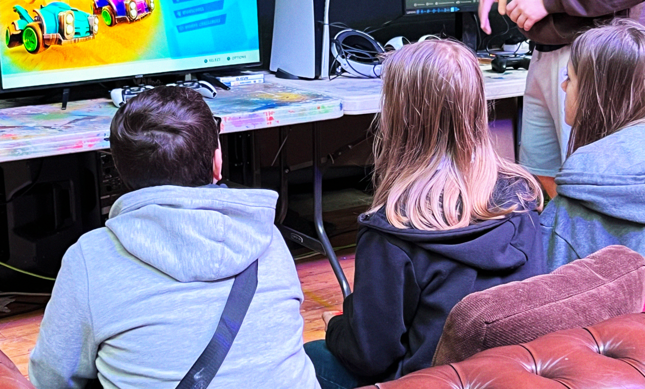 Three young people sit on a brown sofa and play mario kart whilst a man stands off to the right giving instructions for seeing up the game