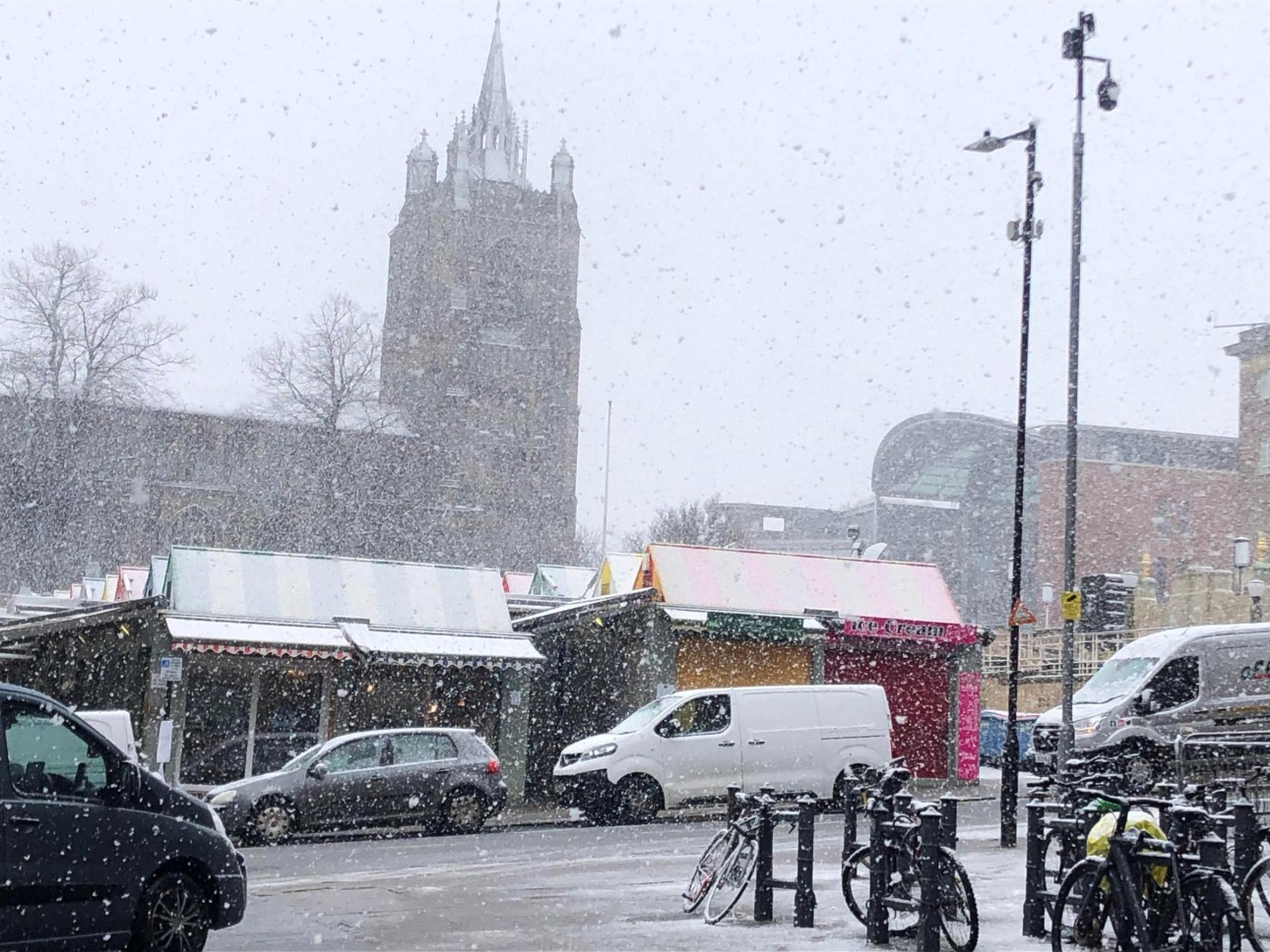Norwich Market blanketed in a layer of sleet as snow continues to fall - the Peter Mancroft Church is visible in the background, above the colorful canopies of the market, and a tiny hint of the Forum is visible to the right