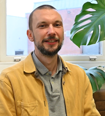 One of our Counsellor's Cai looking at the camera and smiling, whilst seated on a brown leather sofa, with a monteserra plant next to him - and a window behind giving a view of the street and the loading bay of a recently closed Farm Foods.