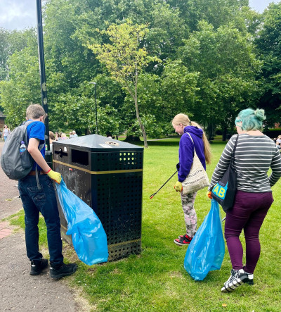 Three young people with litter pickers collecting rubbish in a park and depositing it in blue bin bags
