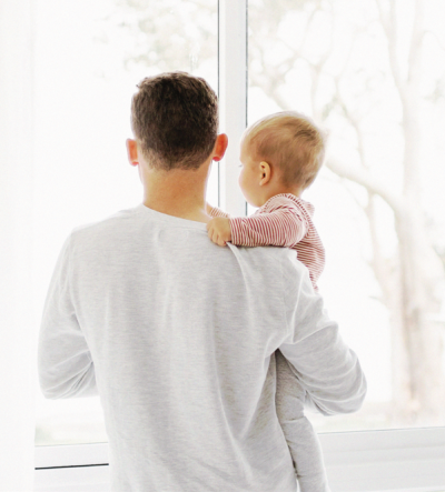 A young father and his infant son stare out of a window, pictured from behind