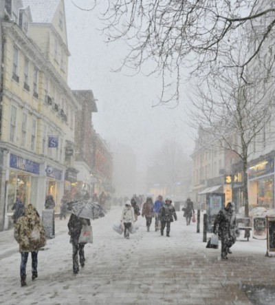 Shoppers walk down Norwich High Street under a driving snow - many are bundled up in winter coats, hoods up, hands stuffed deep inside pockets