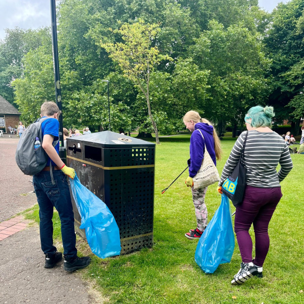 Three young people with litter pickers collecting rubbish in a park and depositing it in blue bin bags