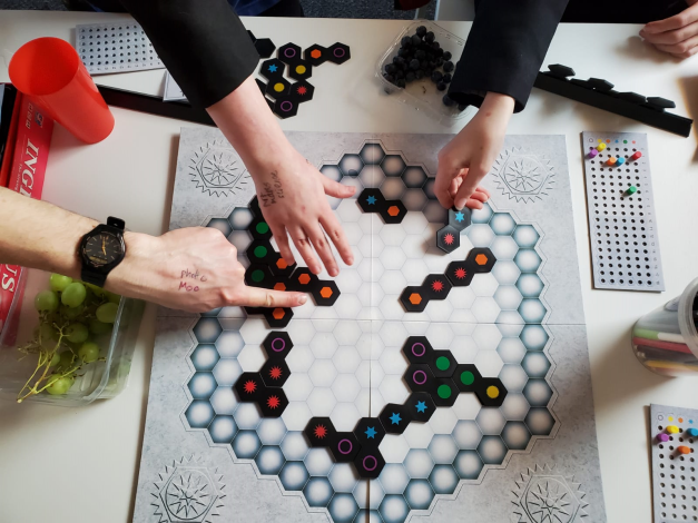 A hexagonal game board with lots of young people's hands reaching across it holding counters