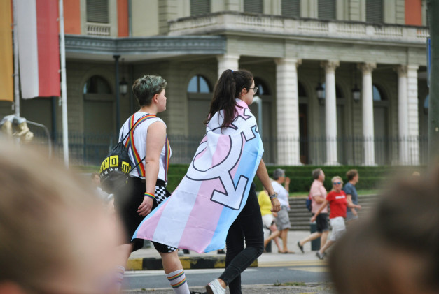 A pair of young protesters walking down a city street in London, one with dyed green hair, the other with a Trans flag as a cape